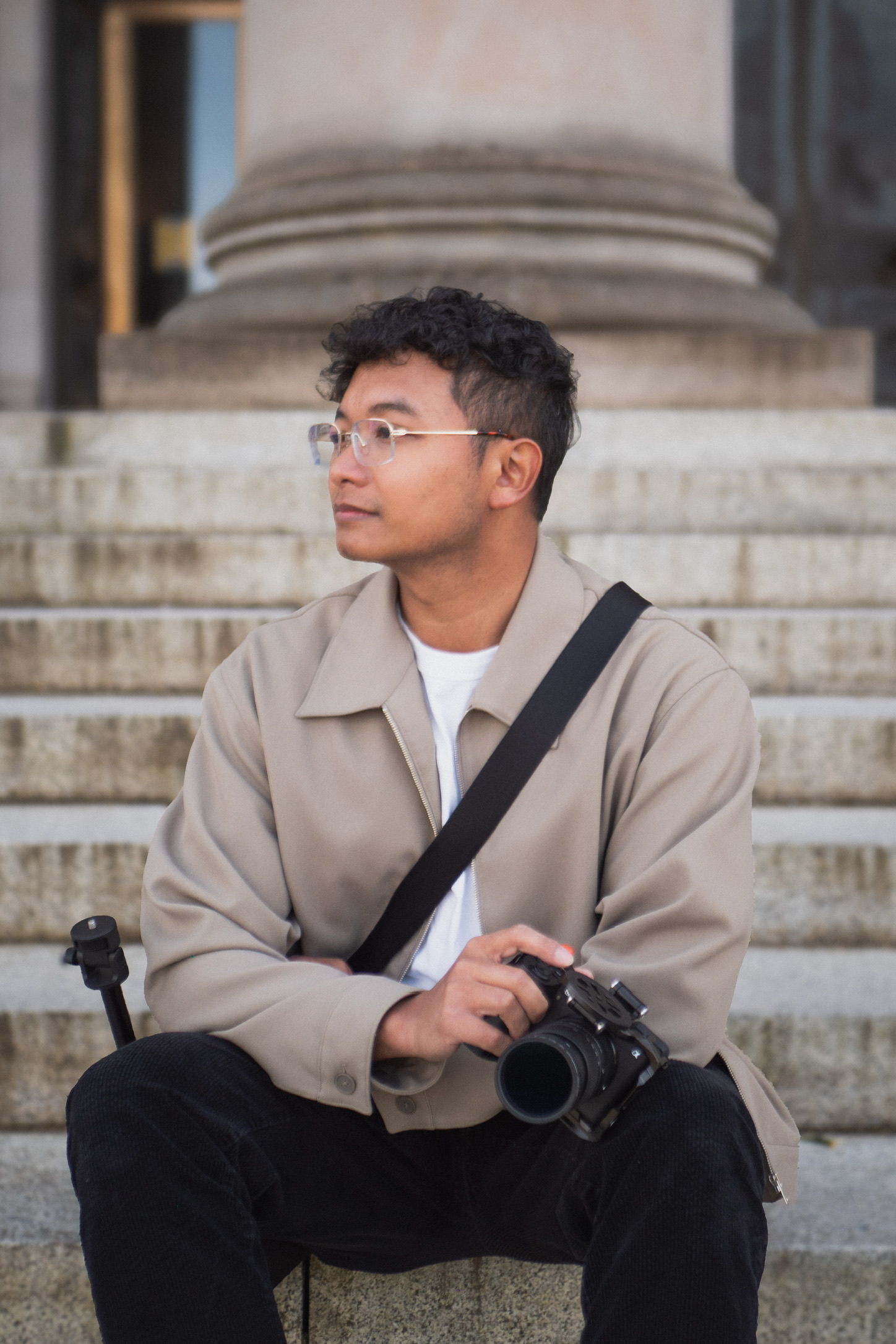 Ricky sitting on stone steps in a beige jacket, holding a camera